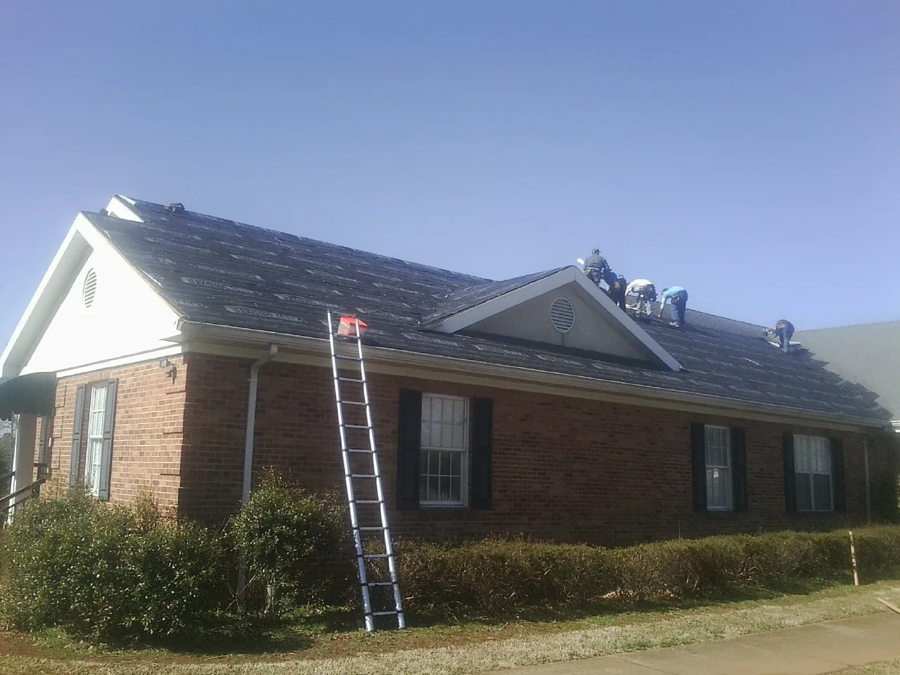 Crew tearing shingles off a brick ranch roof with a ladder set — Emergency Roof Repair work by our crew in Young America