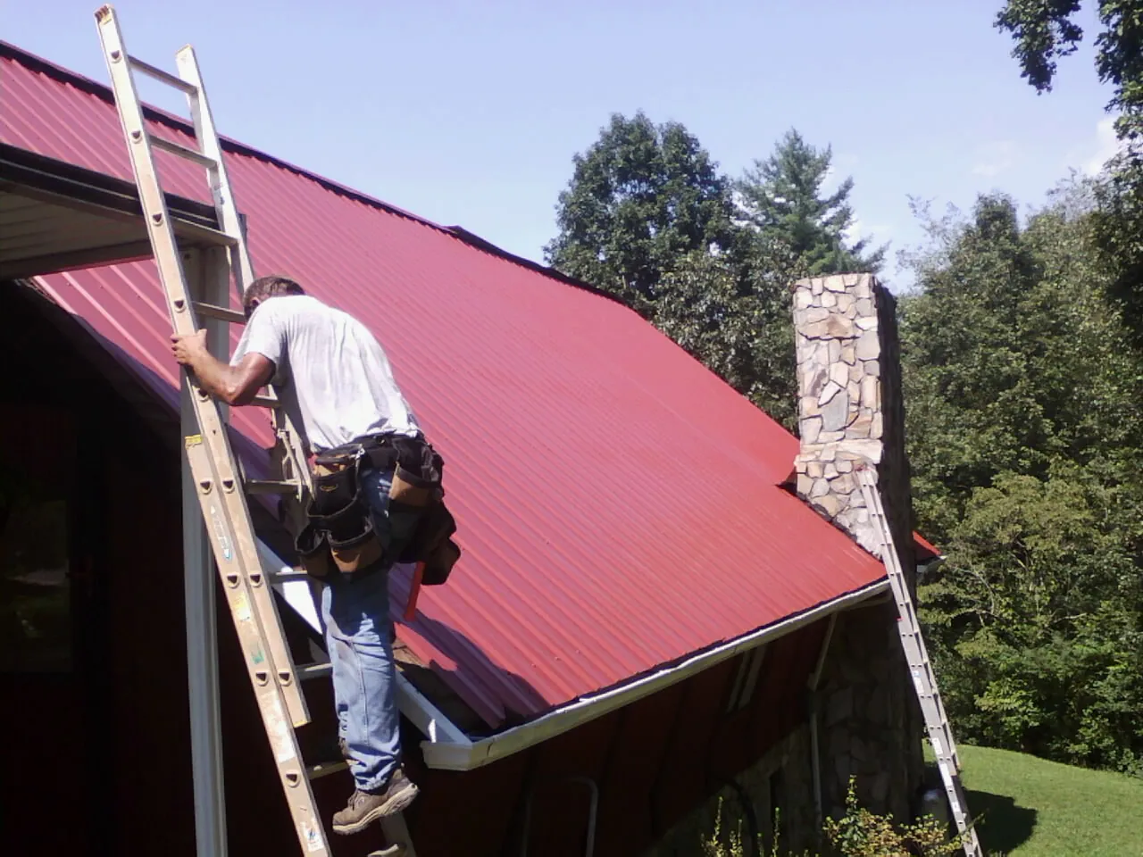 Roofer on a ladder beside a red metal roof and stone chimney — Soffit & Fascia Repair by Roofing Brothers in Young America